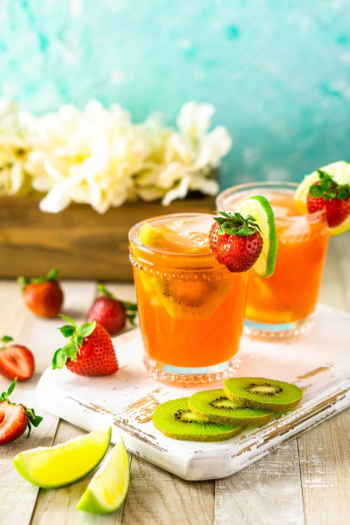 A strawberry-kiwi margarita on a white wooden board with fruit surrounding it and flowers in the background.