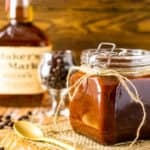 A jar of coffee-bourbon BBQ sauce with a bottle of Maker's Mark and glass of coffee beans in the background.