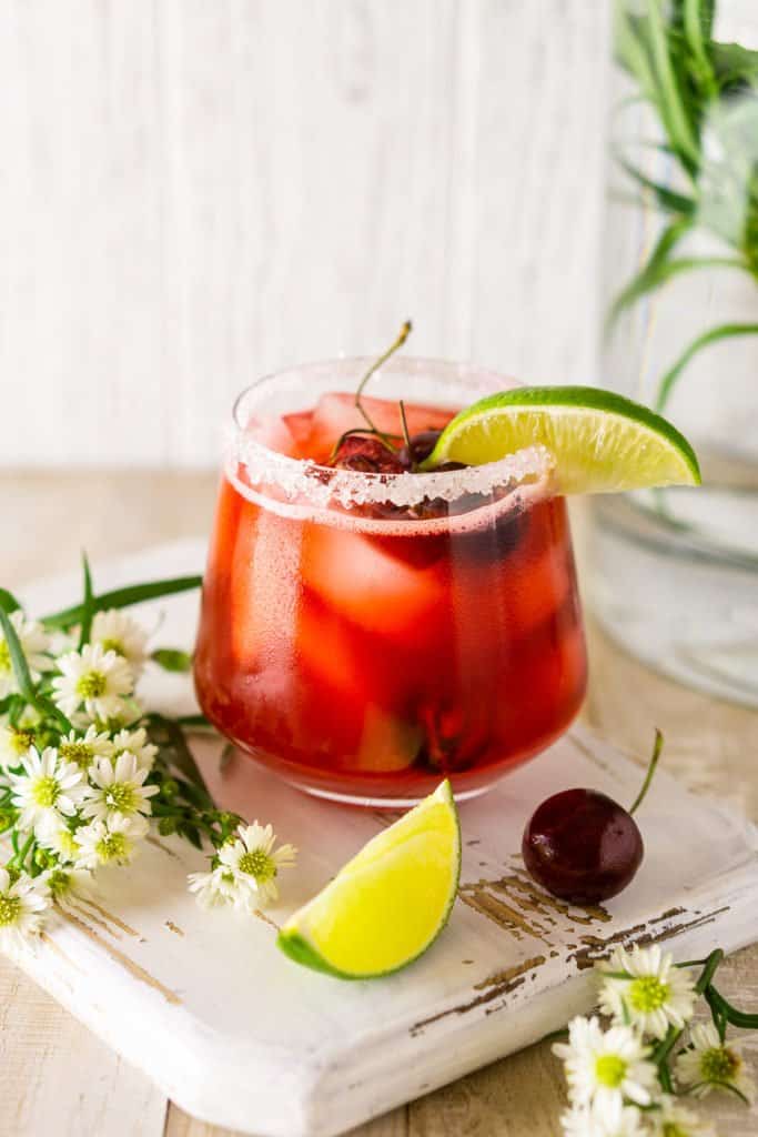 A cherry margarita with a lime slice, cherry and bundle of flowers on the side.