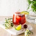 A fresh cherry margarita on a white wooden board with white flowers in a vase in the background.