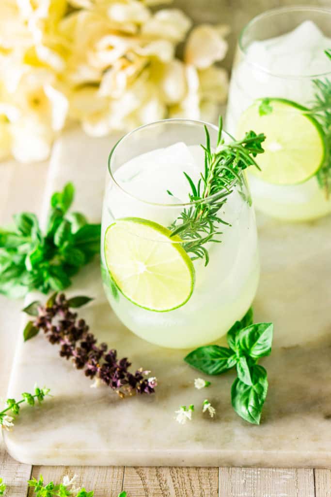 Looking down at a gin and tonic with fresh herbs from the garden around it on a white marble board.