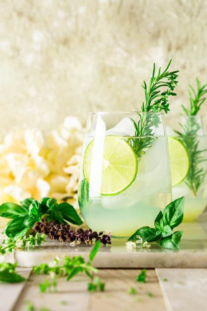 A garden gin and tonic with a rosemary sprig garnish and fresh herbs surrounding it.