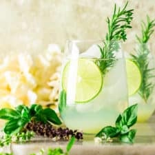 A garden gin and tonic with a rosemary sprig garnish and fresh herbs surrounding it.