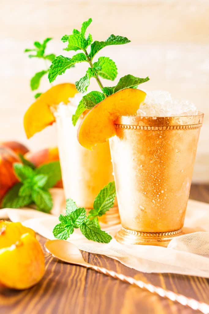 Two brown sugar-peach mint juleps with fresh mint sprigs and peach slices on the side.
