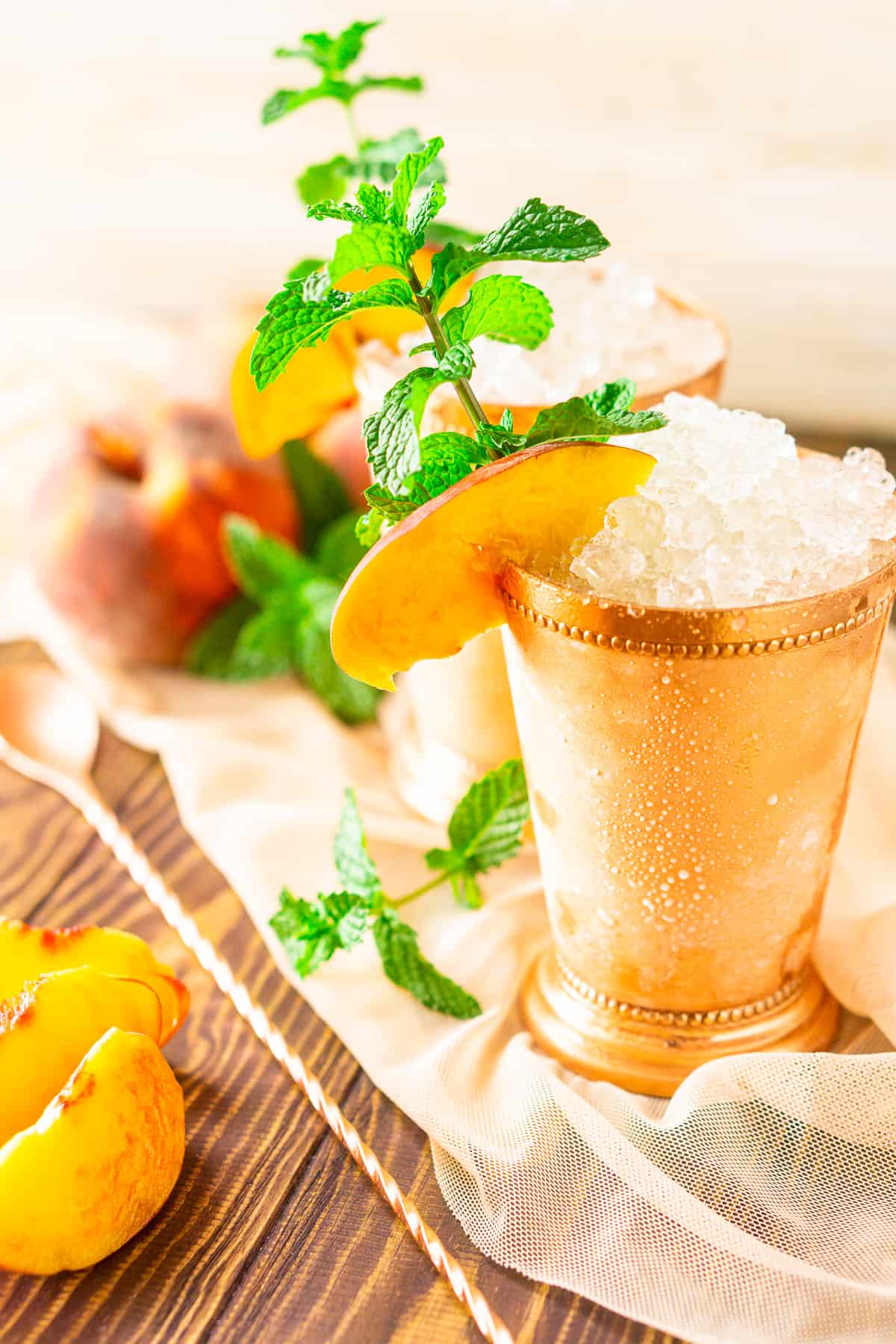 The peach mint juleps with a bunch of whole peaches in the background on sheer fabric.