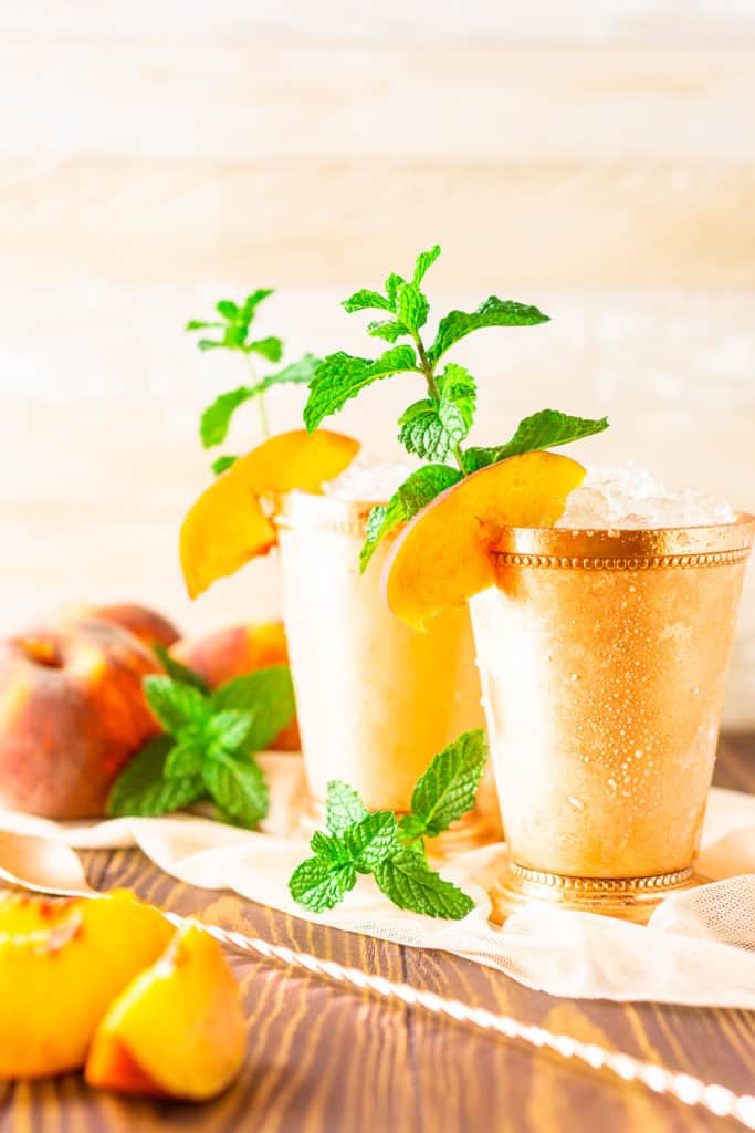 Two brown sugar-peach mint juleps with a bar spoon and peach slices next to them.