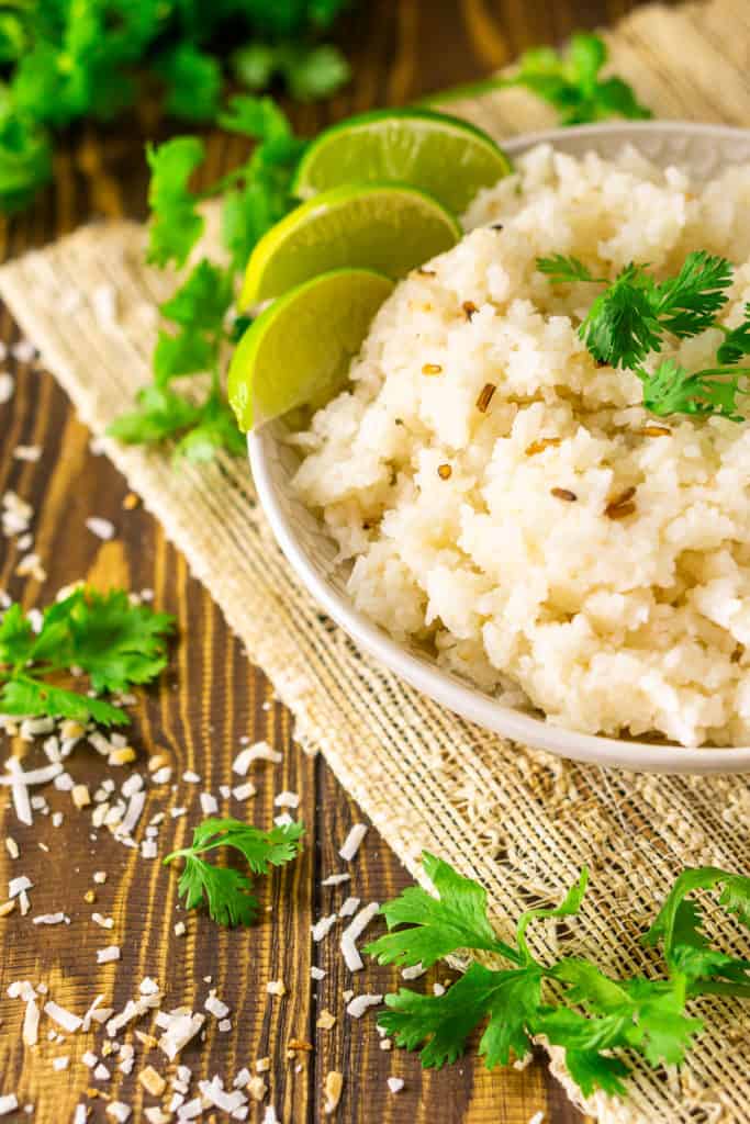 A bowl of tropical coconut-lime rice on a placemat with toasted coconut and cilantro around it and two spoons in the background.