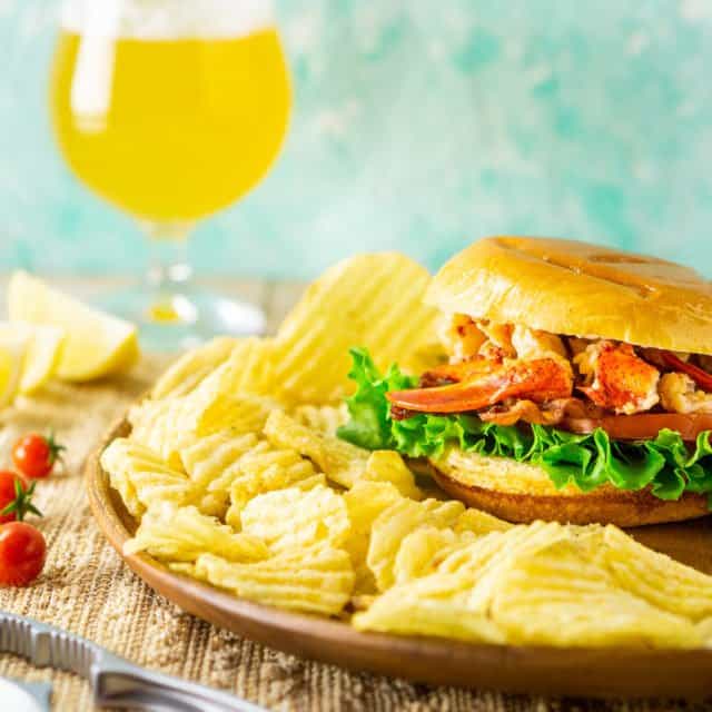 A Connecticut-style lobster BLT with chips around it, a lobster claw cracker on the side and a beer in the background.