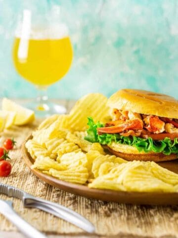 A Connecticut-style lobster BLT with chips around it, a lobster claw cracker on the side and a beer in the background.