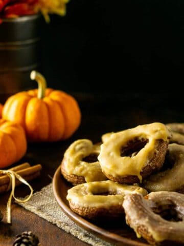 A plate of pumpkin cruller donuts on a wooden plate with pumpkins and cinnamon sticks to the left.