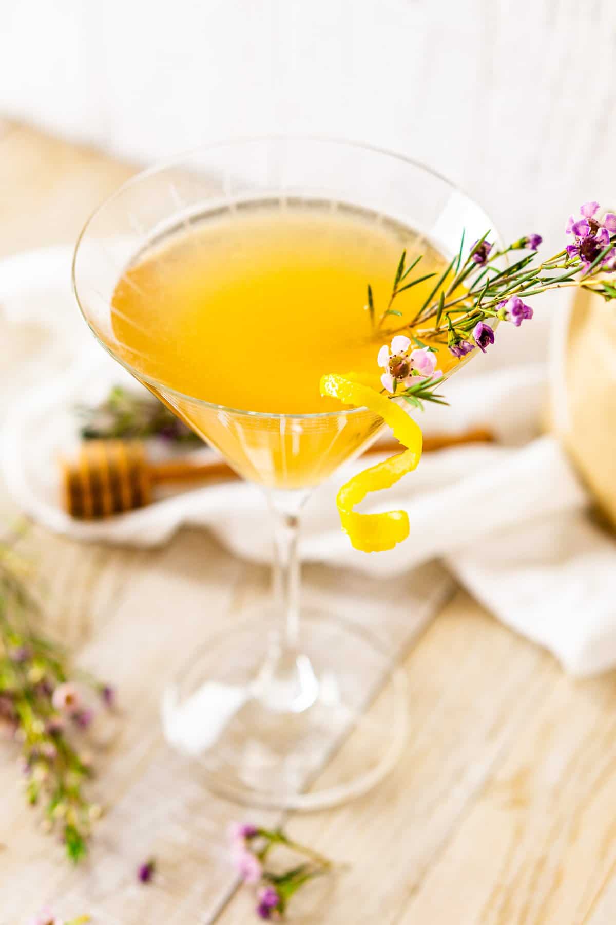 A side view and looking down onto a lavender bee's knees with a lemon twist and floral garnishes on a white wooden board.