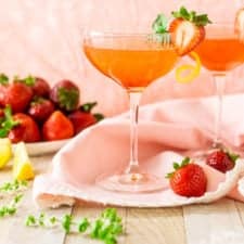 Two strawberry-basil limoncello martinis on a pink napkin with strawberries and basil flowers around it.