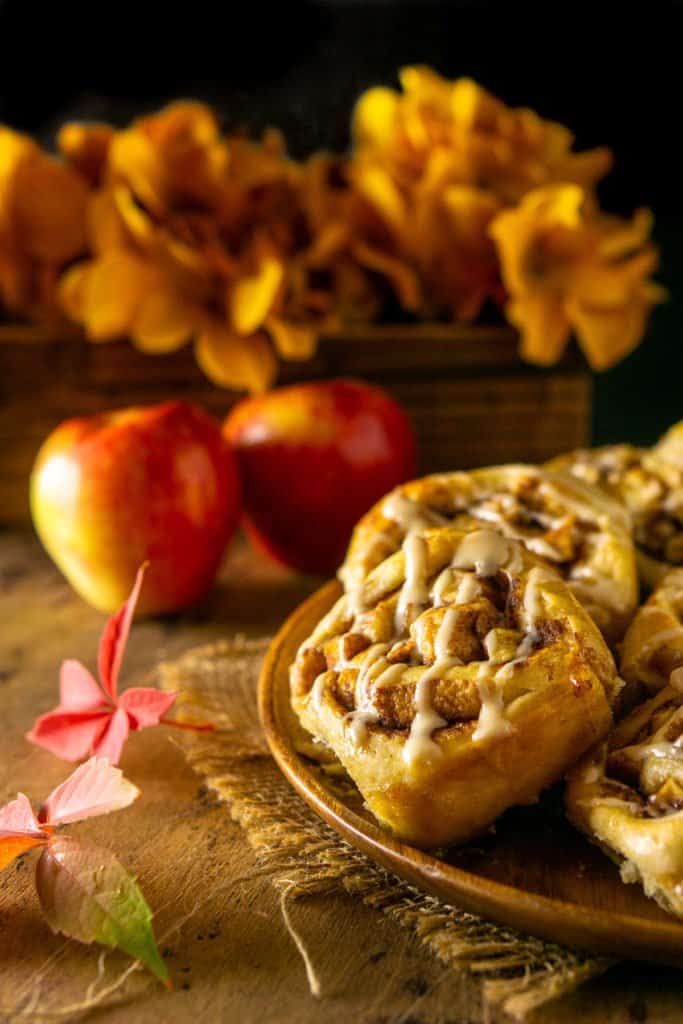 A wooden plate of apple cinnamon rolls with apple and flowers in the background.