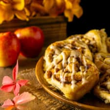 A plate of apple cinnamon rolls on burlap with fall leaves and flowers in the background.