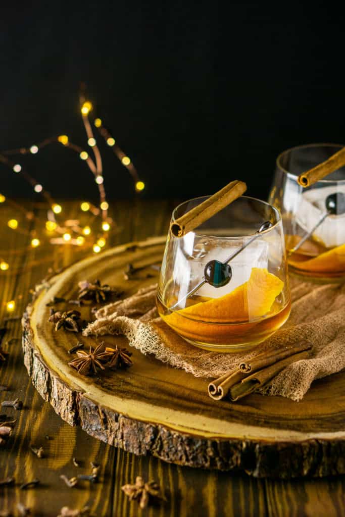 Two chai old fashioned cocktails on a wooden platter with spices on the side and lights in the background.