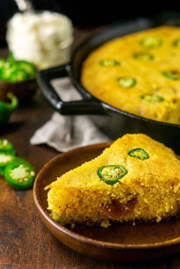 A slice of jalapeno popper cornbread on a small wooden plate in front of the cast-iron skillet.