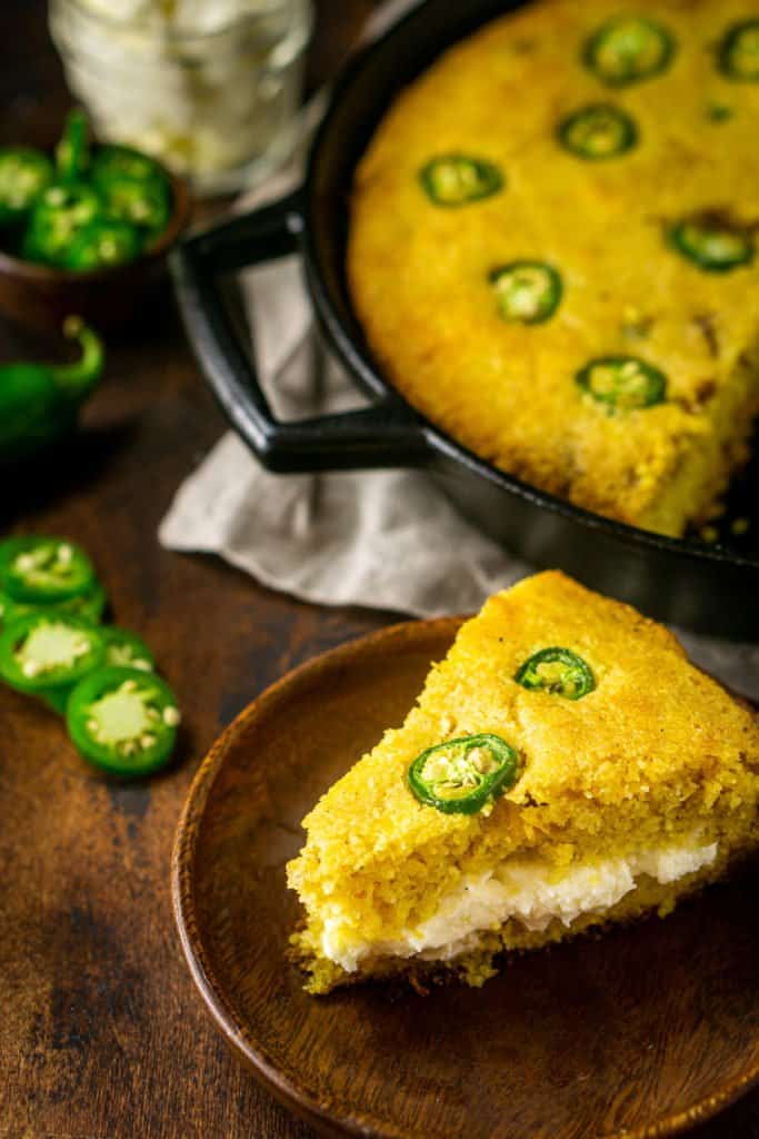 Looking down on a slice of jalapeno popper cornbread filled with the whipped cream cheese butter with a skillet of cornbread behind the slice and jalapenos on the side.