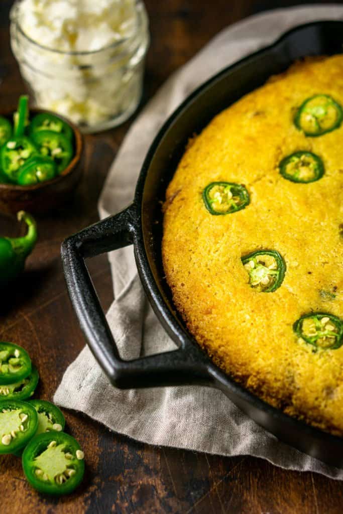 The jalapeno popper cornbread in a cast-iron skillet with a mason jar of whipped cream cheese butter behind it and jalapenos on the side.
