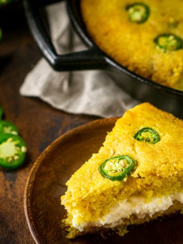 Looking down on a slice of jalapeno popper cornbread filled with the whipped cream cheese butter with a skillet of cornbread behind the slice and jalapenos on the side.