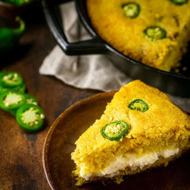 Looking down on a slice of jalapeno popper cornbread filled with the whipped cream cheese butter with a skillet of cornbread behind the slice and jalapenos on the side.