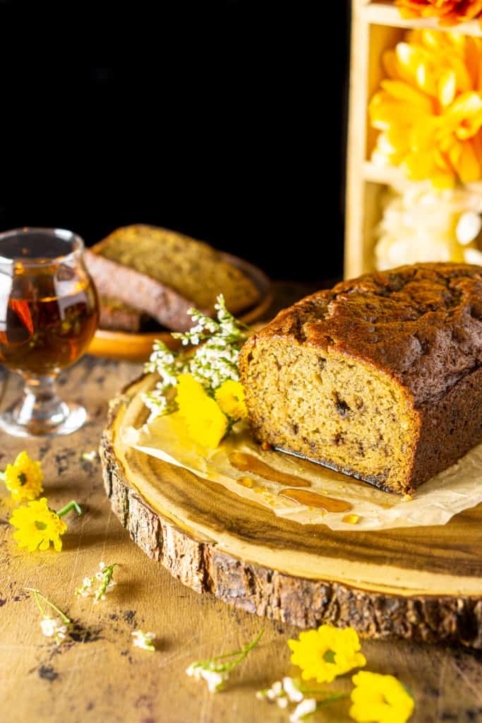 A loaf of this boozy banana bread with a glass of bourbon behind it and flowers to the side.