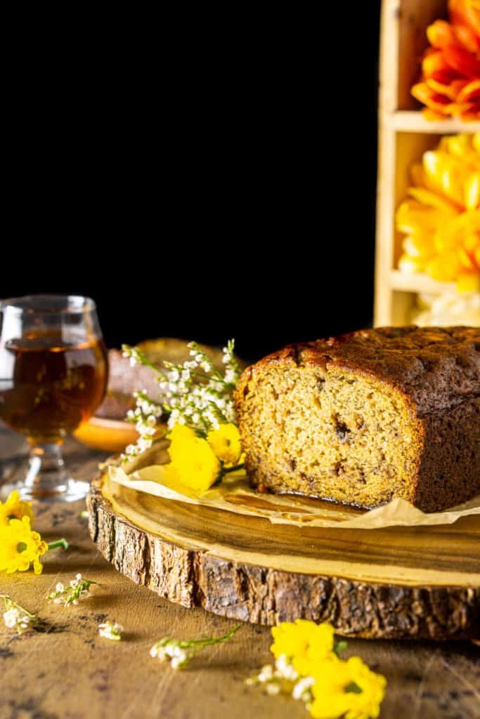 A cut loaf of maple-bourbon banana bread on a wooden platter with a glass of bourbon to the side and flowers surrounding it.