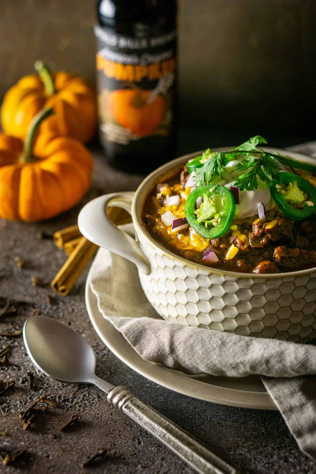 A bowl of pumpkin and chorizo chili with two small pumpkins and a pumpkin beer in the background with a spoon to the side.