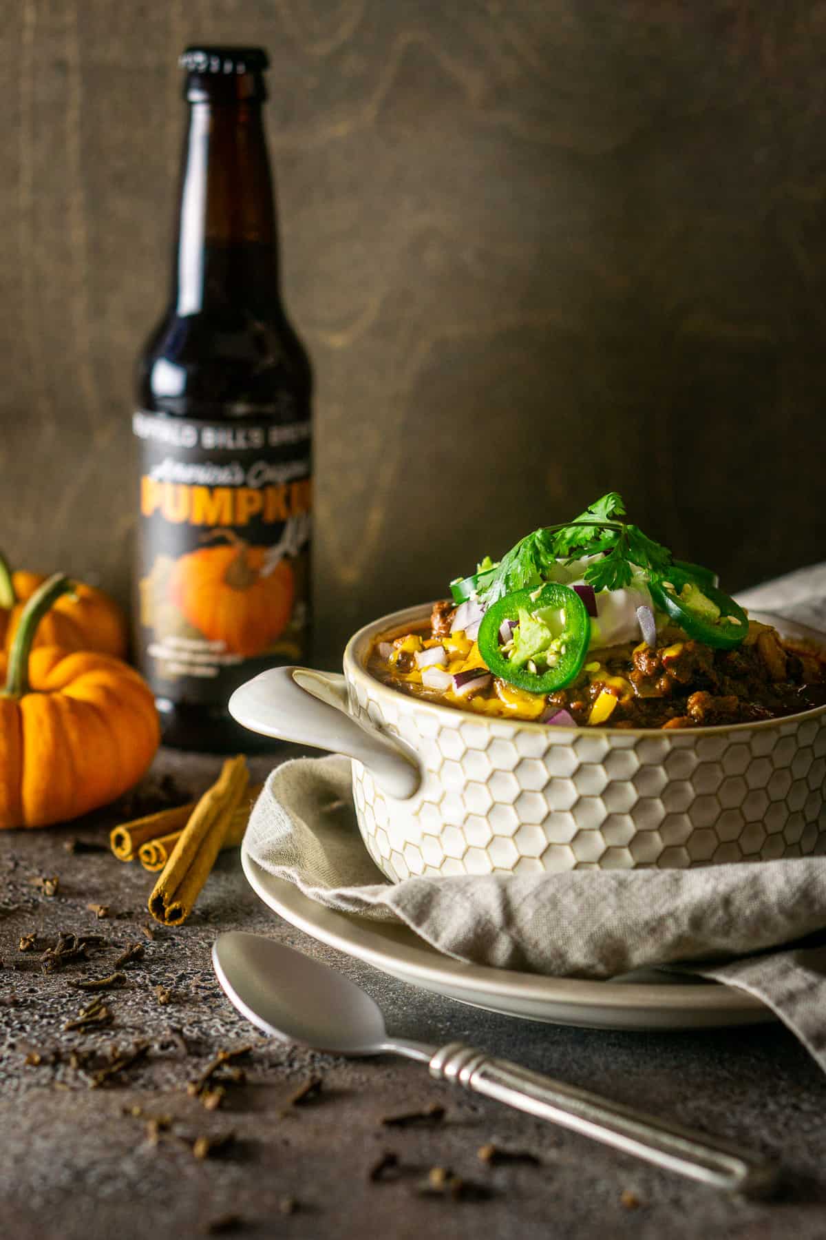 A bowl of pumpkin and chorizo chili with cloves and cinnamon sticks to the side and a pumpkin beer in the background.
