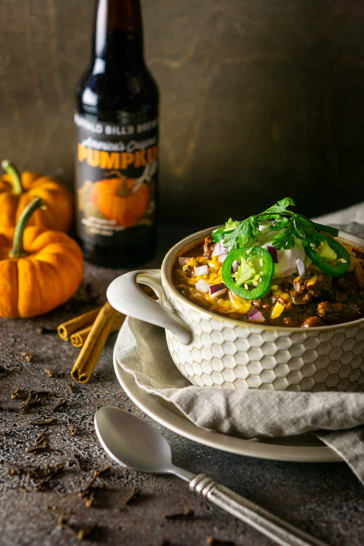 The pumpkin and chorizo chili on a small plate with a clothe napkin and a beer in the background with pumpkins.