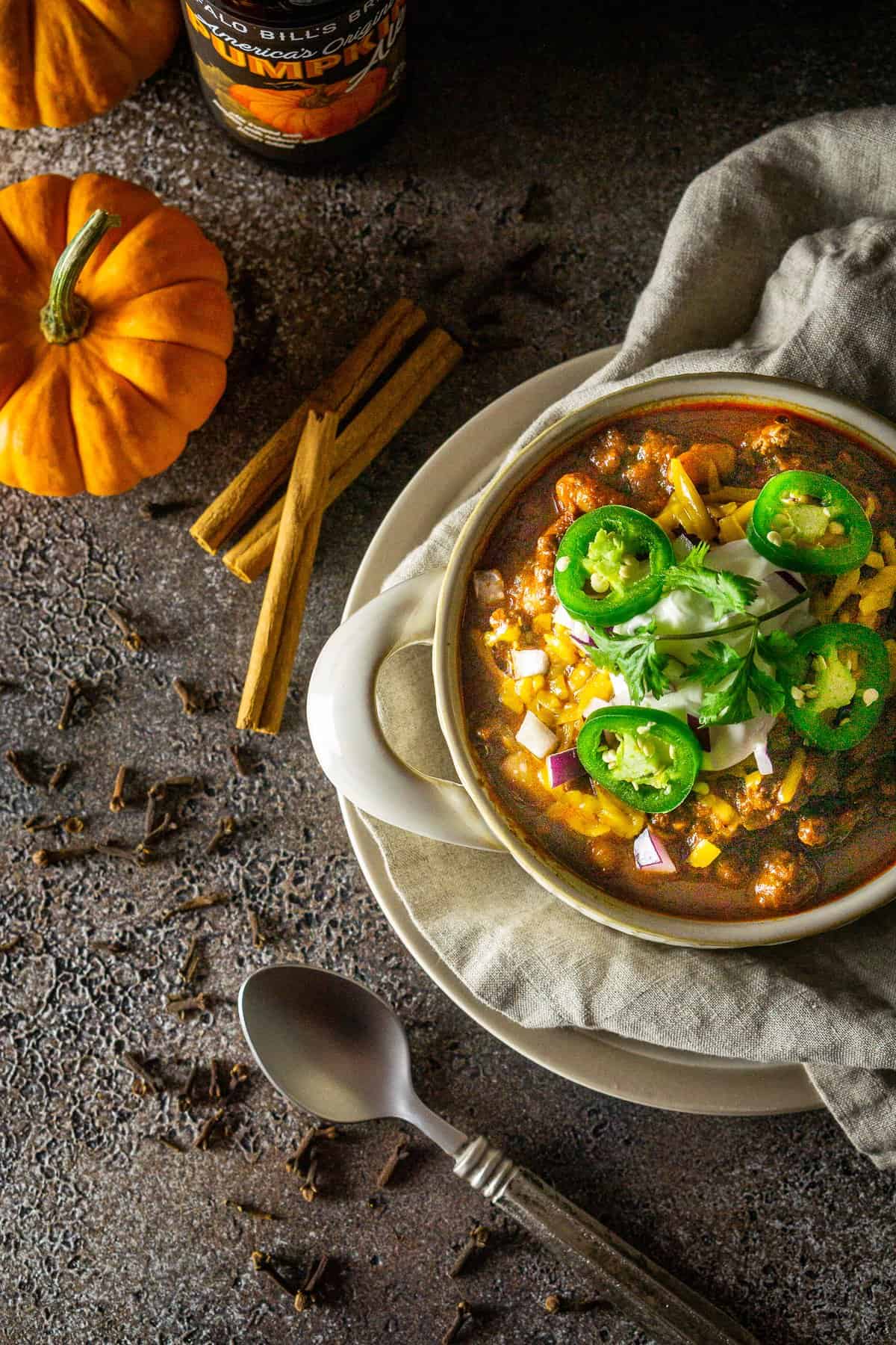 An aerial view of the pumpkin and chorizo chili with spices, a spoon, pumpkin and a beer to the side.