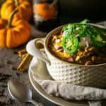 A bowl of pumpkin and chorizo chili with two small pumpkins and a pumpkin beer in the background with a spoon to the side.