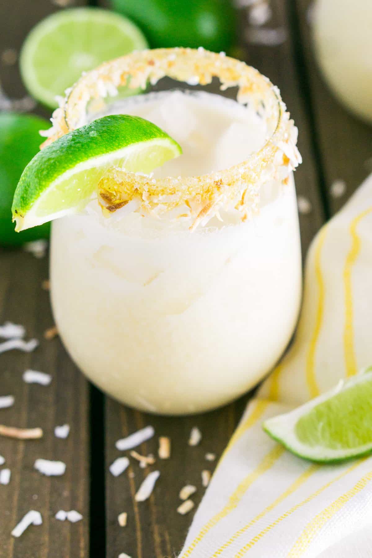 A coconut margarita with a yellow napkin and lime slices on the side with toasted coconut scattered around it.