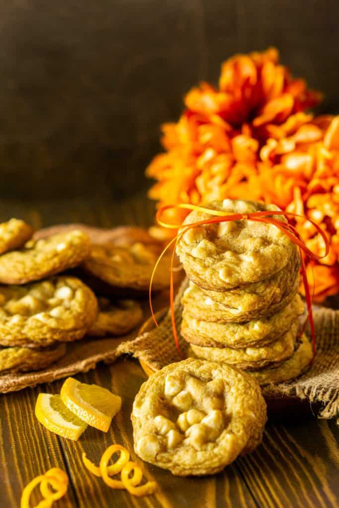 A stack of ginger-maple cookies with a bunch of orange flowers behind it and a couple orange zest twists on the side.