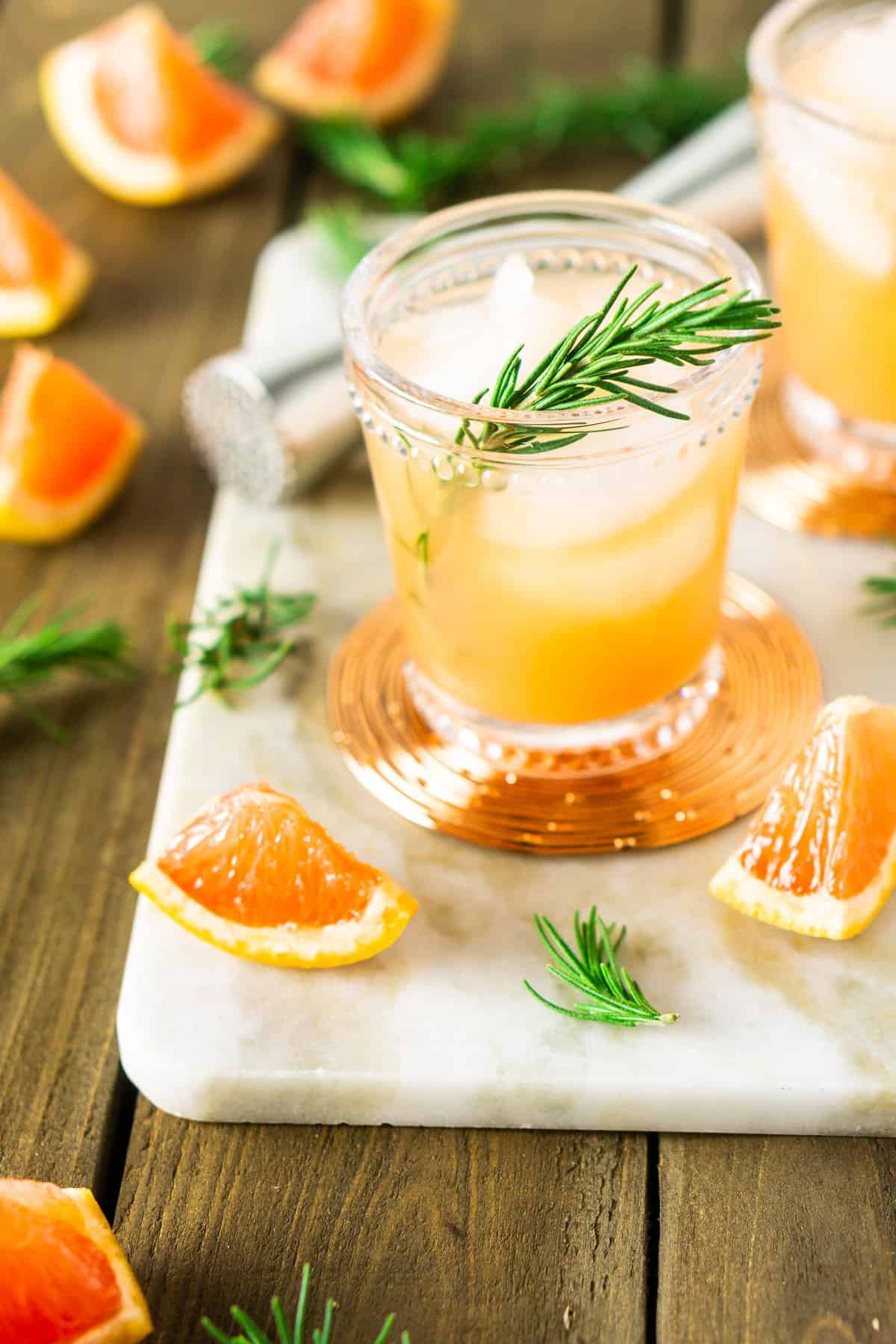 A rosemary-grapefruit spritzer on a white marble tray with bar tools in the background and fresh fruit around it.