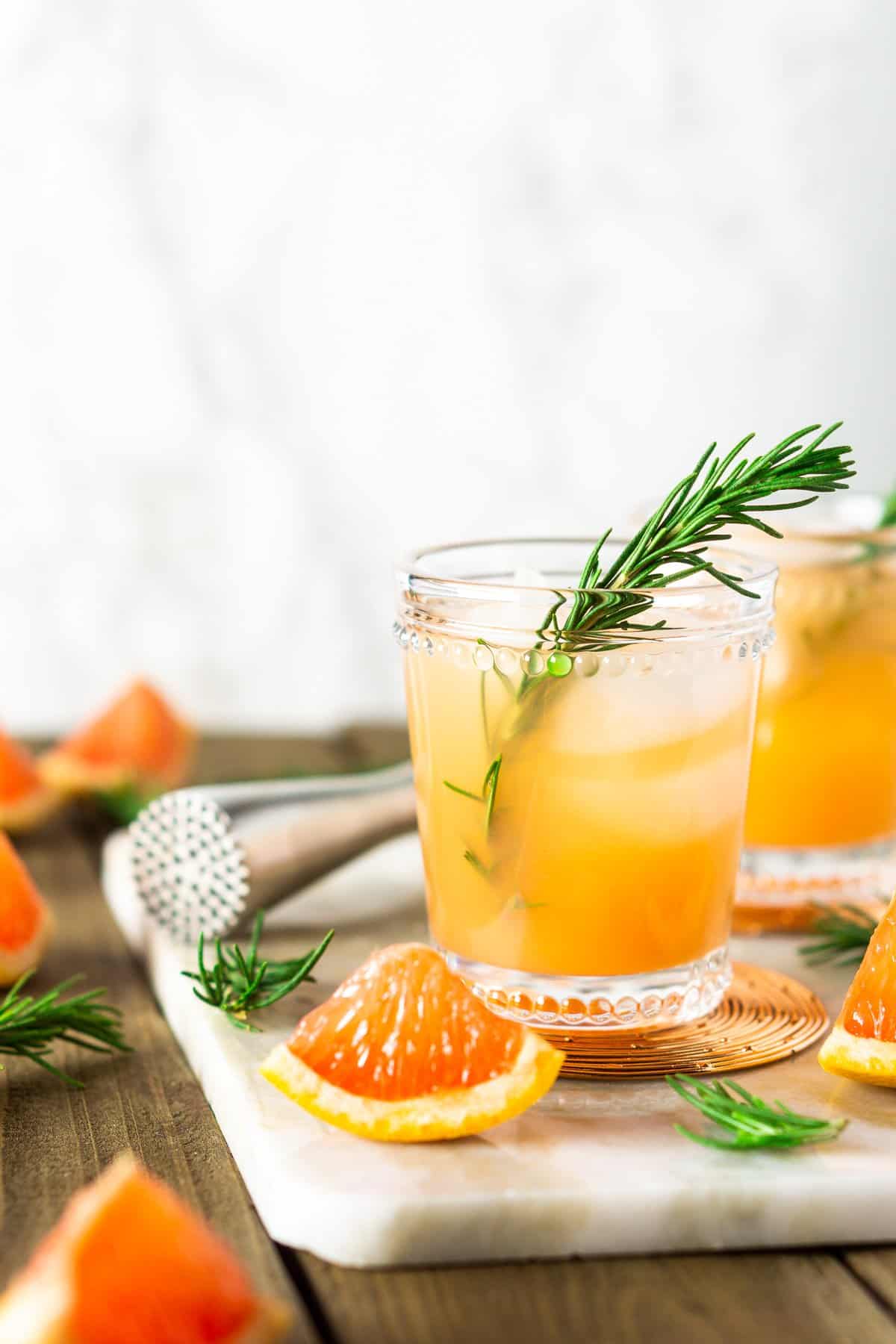 Two rosemary-grapefruit spritzers on a white tray against a marble background with fruit and bar tools around them.