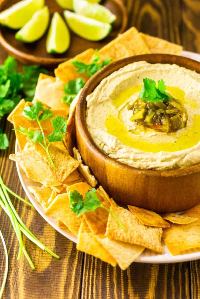 A bowl of creamy Hatch green chile hummus with pita chips on the side, lime slices in the background and pieces of fresh cilantro.