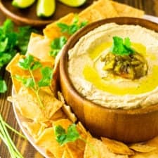 A bowl of creamy Hatch green chile hummus with pita chips on the side, lime slices in the background and pieces of fresh cilantro.