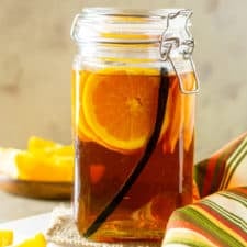 A jar of vanilla bean and orange infused bourbon with orange slices and a cloth napkin to the side.