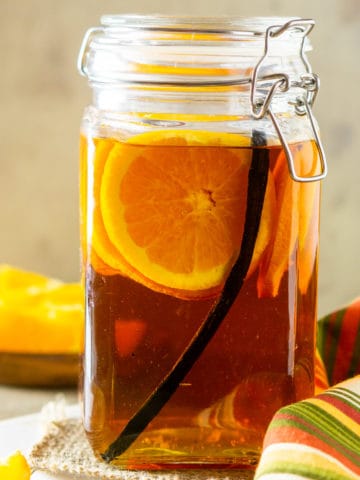 A jar of vanilla bean and orange infused bourbon with orange slices and a cloth napkin to the side.