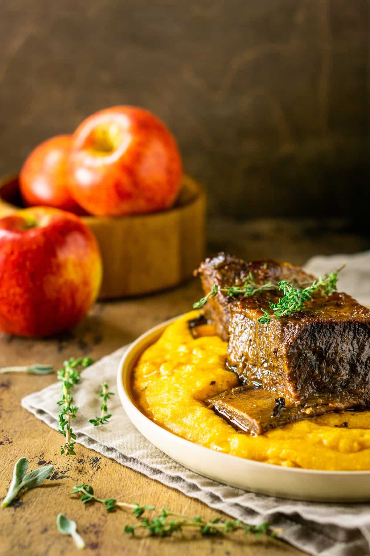 A plate of polenta with a braised apple cider short rib with fresh herbs around it.