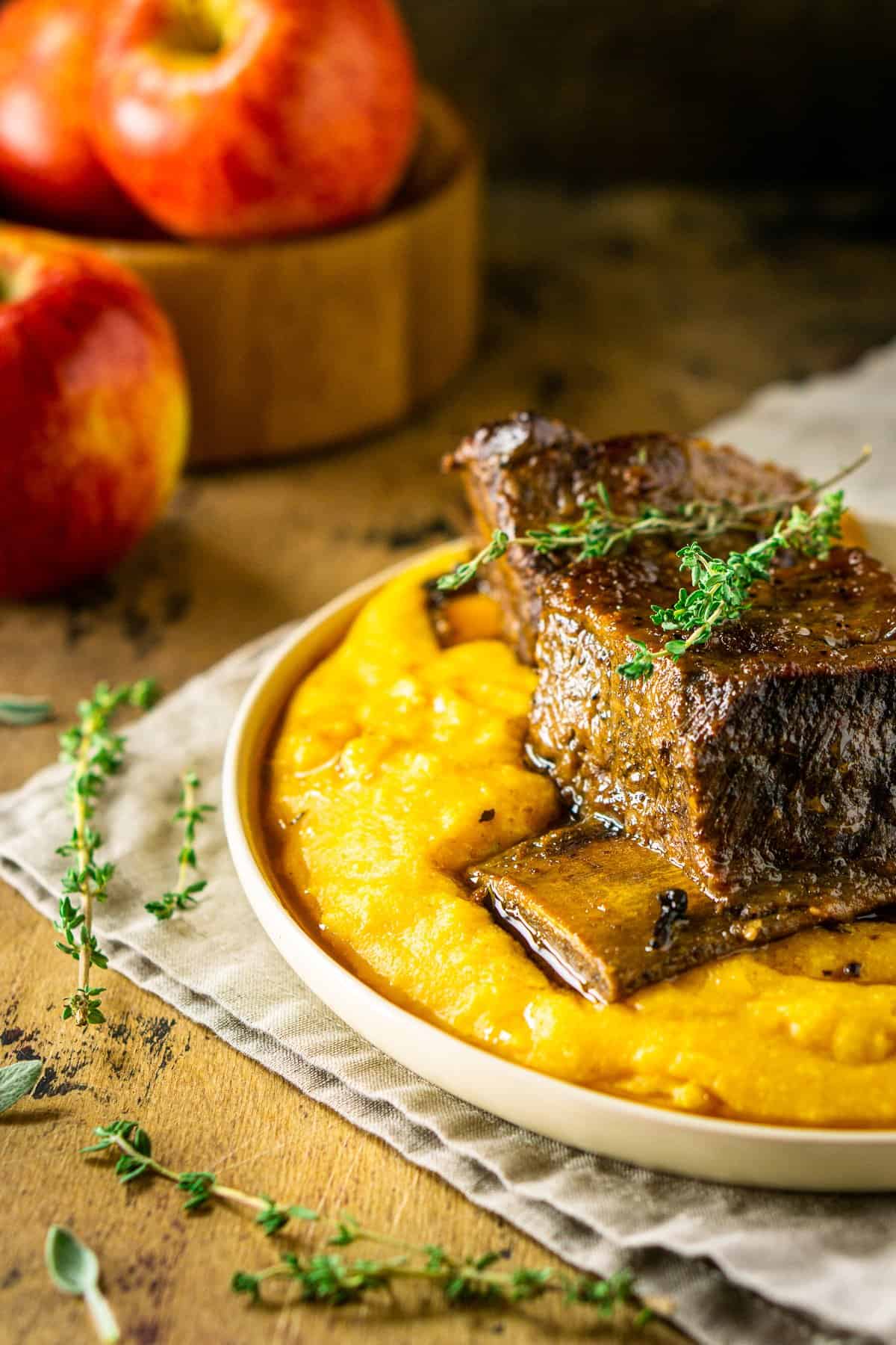 Two braised apple cider short ribs on polenta with fresh herbs to the side and apples in the background.