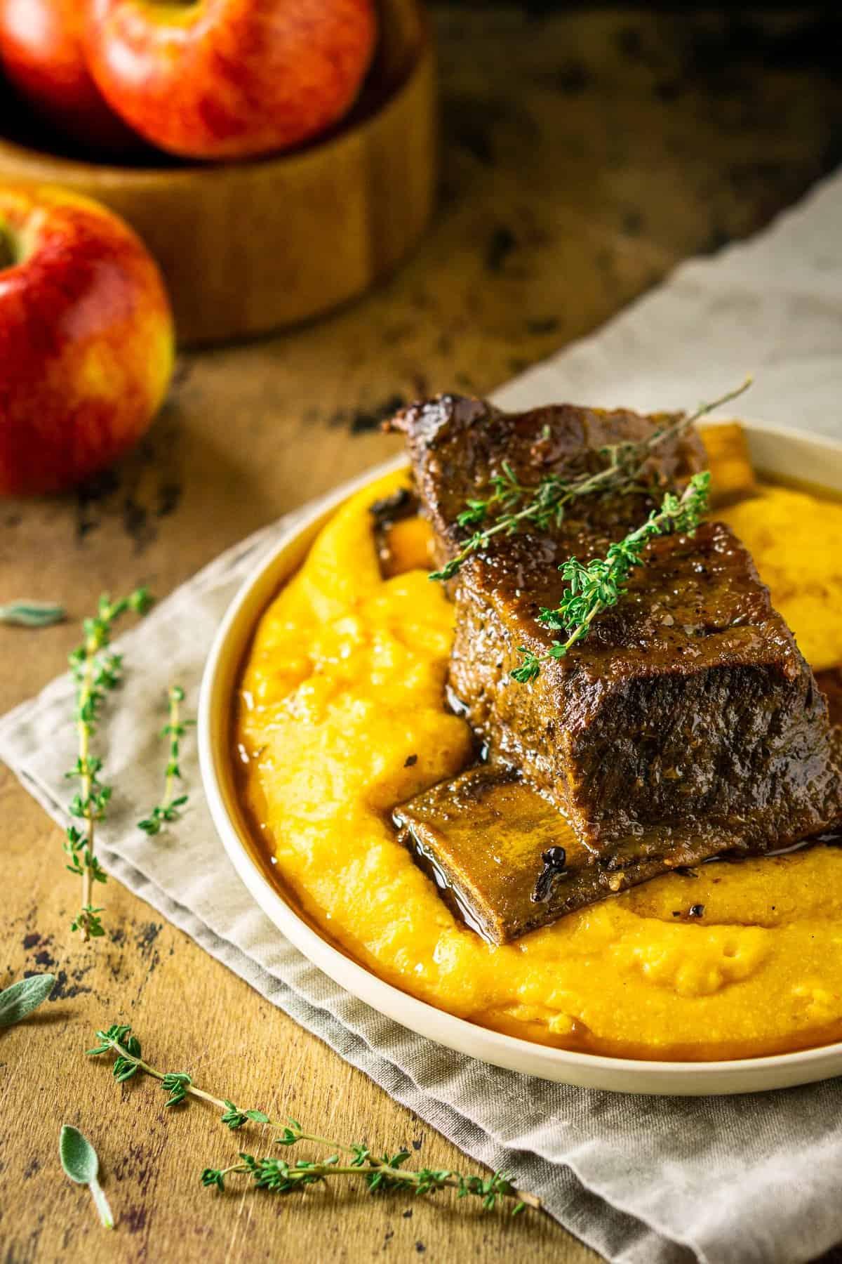 A side view of two apple cider short ribs with a wooden bowl of apples behind it.