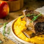 Two braised apple cider short ribs on polenta with fresh herbs to the side and apples in the background.