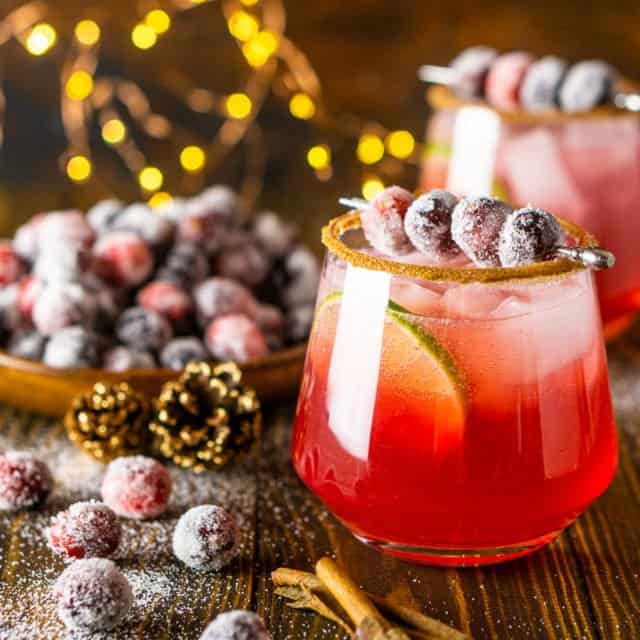 A sparkling cranberry-ginger margarita with sugared cranberries to the side and holiday lights behind it.