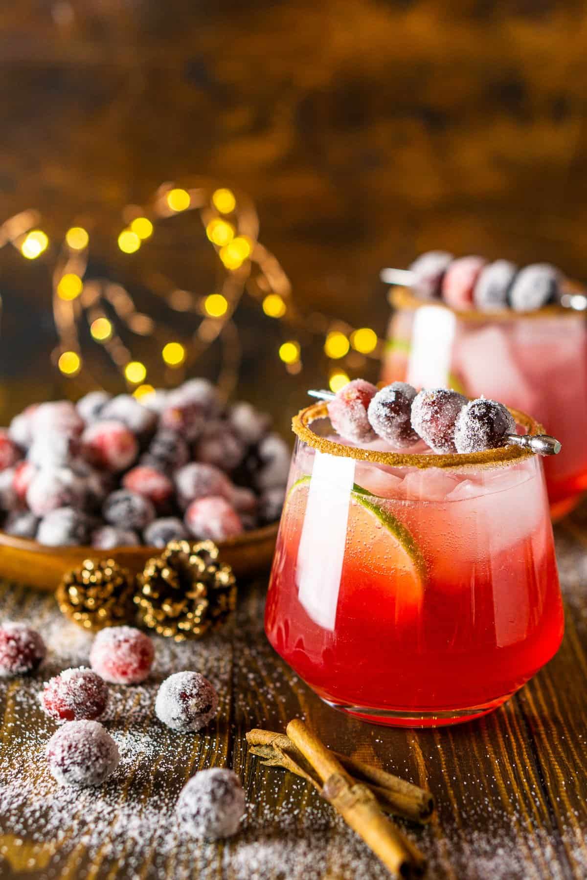 A sparkling cranberry-ginger margarita with sugared cranberries to the side and holiday lights behind it.