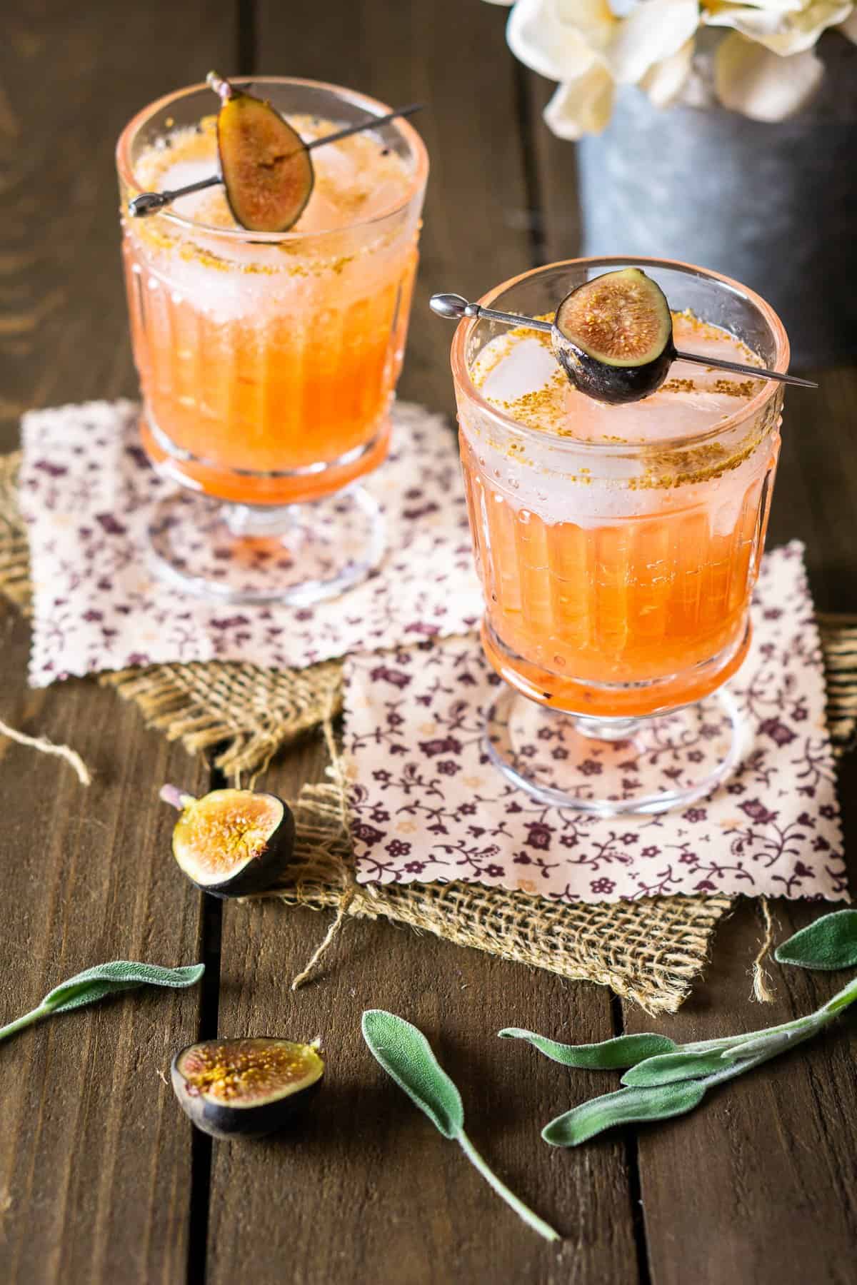 Two sage-fig cocktails on burlap and fabric with white flowers behind them.