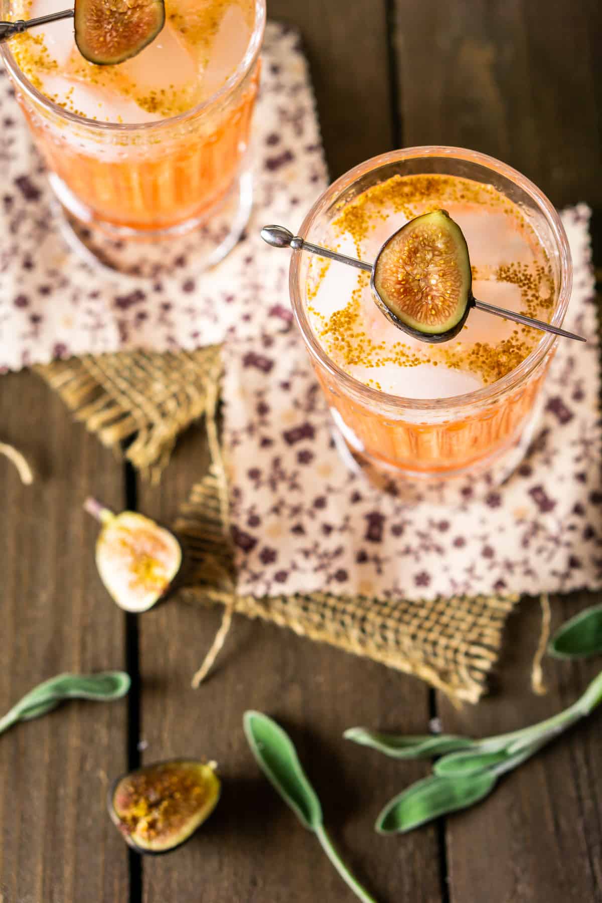 Two sage and fig cocktails from an aerial view on burlap with sliced figs on the side.