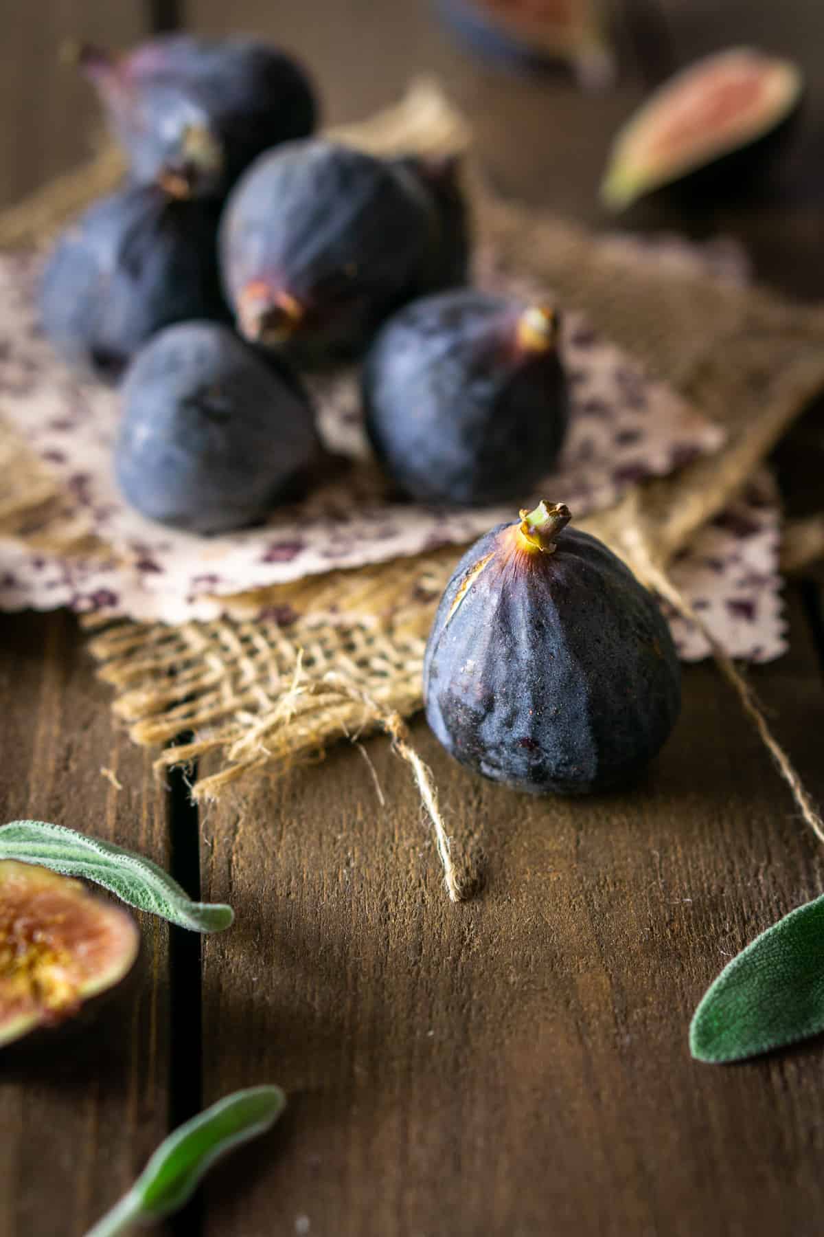 A pile of fresh black mission figs on burlap with fresh sage around them.