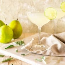 Two pear-sage gimlet cocktails on a marble board with fresh sage around it and pears in the background.