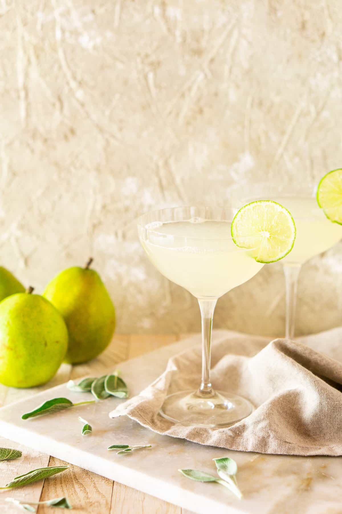 Two pear-sage gimlet cocktails on a marble board with fresh sage around it and pears in the background.